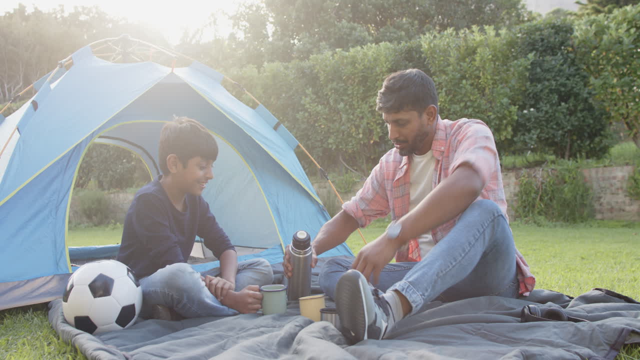 Camping outdoors, Indian father pouring drink for son near tent and soccer ball