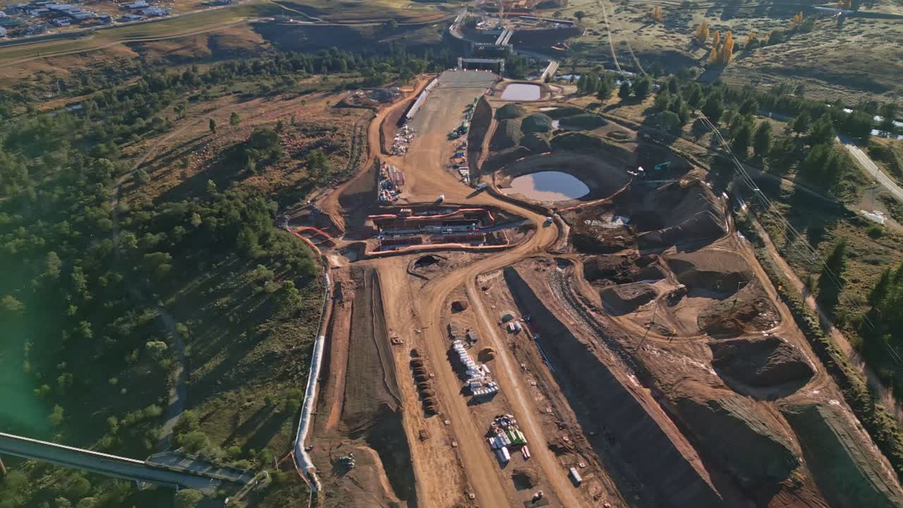 Sweeping drone shot around highway construction with river below and future road path visible, enhanced by a soft lens flare.