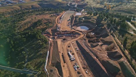 sweeping drone shot around highway construction with river below and future road path visible, enhanced by a soft lens flare.