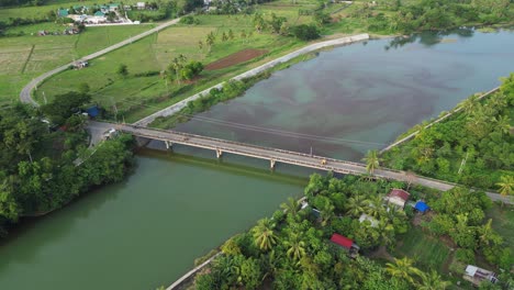 Aerial-overview-of-vibrant-river-and-bridge-next-to-quaint-Philippine-village-in-tropical-island-Catanduanes,-Bicol