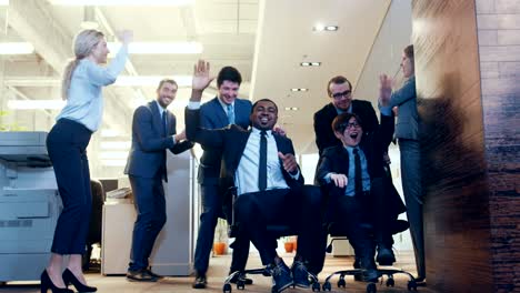 office chair race funny multi ethnic businessmen ride chairs in the corporate building hallway. colleagues cheer, applaud and high-five. celebrate closing of the deal.