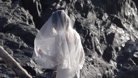 mujer elegante con velo de novia blanco y vestido durante una sesión de fotos en la playa rocosa