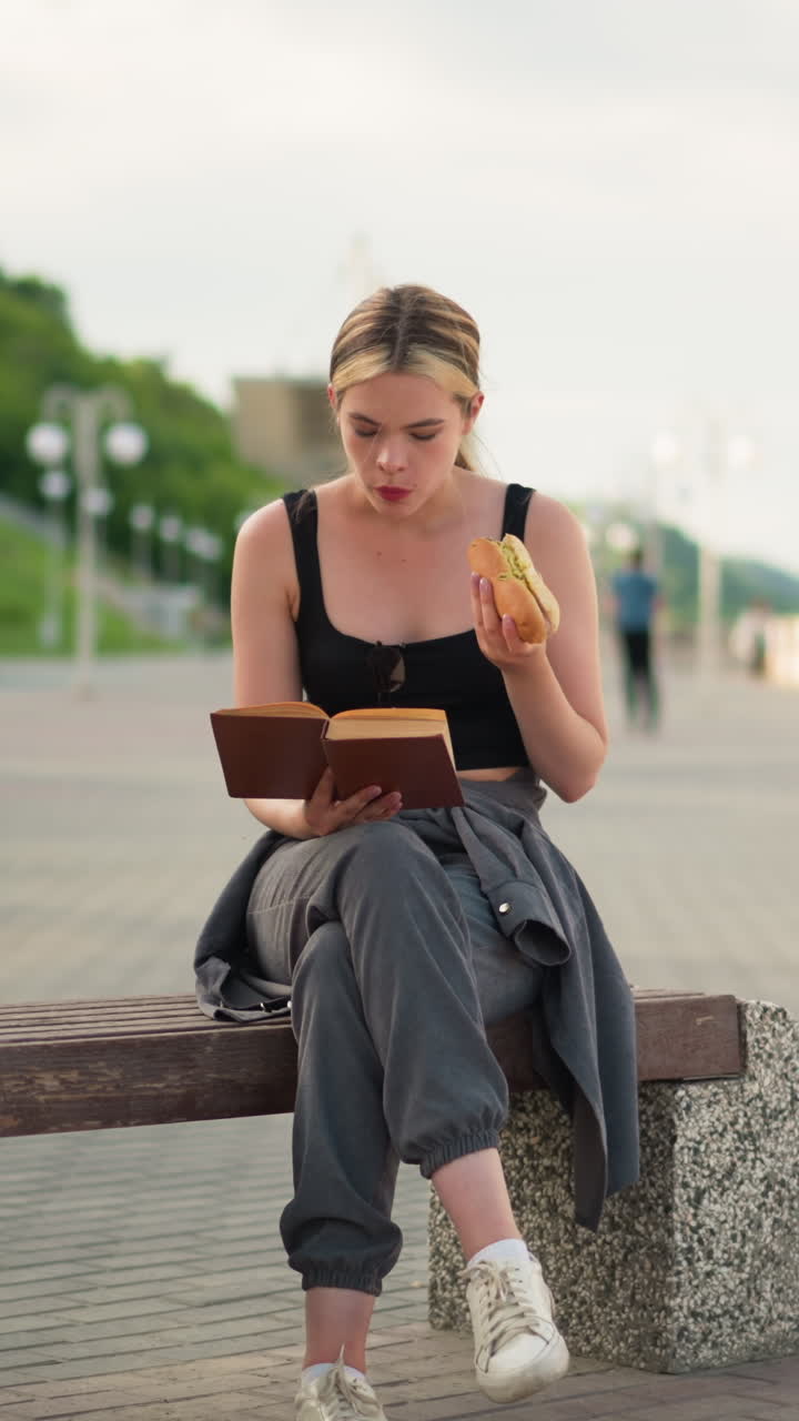 Woman seated outdoors with crossed legs, holding a book in one hand while eating snacks and reading, background features lamp post and greenery hill in a peaceful park setting