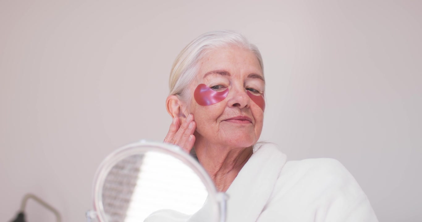 Anziana donna in accappatoio che inizia la routine di cura della pelle alla toeletta, applicando patch occhi rosa mentre si guarda allo specchio.