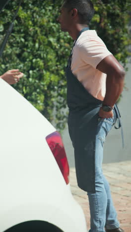 vertical video: man in apron talking to woman outside, standing near car and greenery