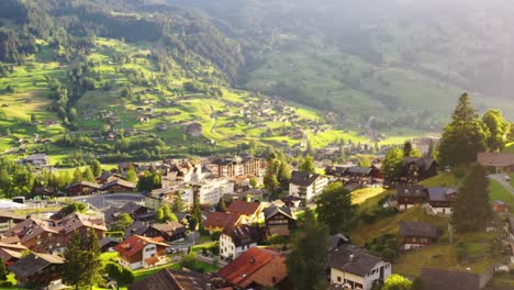 aerial drone view of dreamy swiss mountain village grindelwald, wooden chalet cabins scattered upon alpine green rolling hills and meadows between majestic peaks at sunset golden light, switzerland