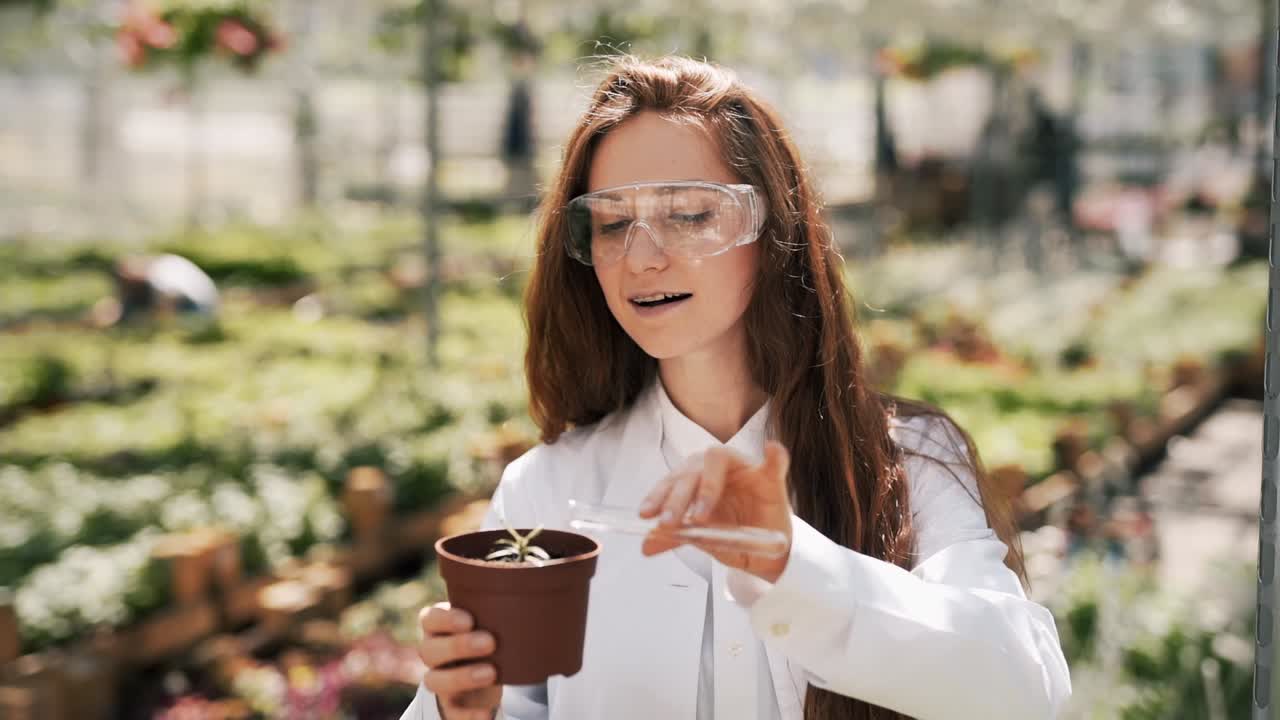 woman in a white coat and goggles pouring a test tube with water a pot with a plant