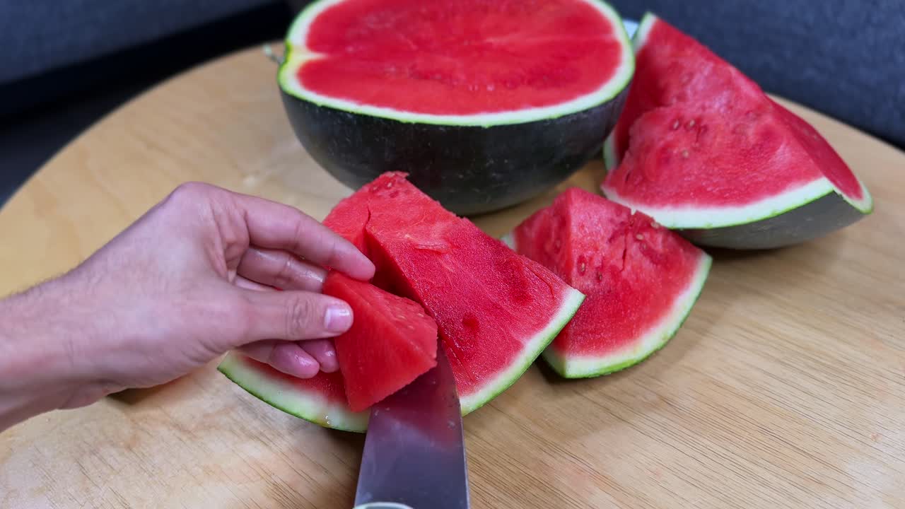 Close-up of a hand slicing a juicy watermelon with a knife on a wooden table, ideal for food, health, nutrition, summer refreshment, and lifestyle projects