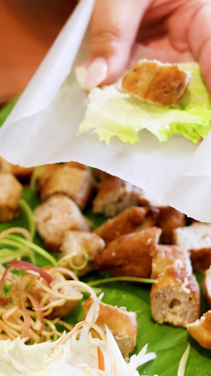 Bidikan jarak dekat tangan yang meletakkan daging babi panggang di atas kertas nasi dengan selada dan rempah-rempah.