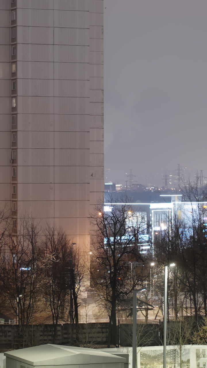 Night view of a tall urban apartment building with bare trees and city lights in the background