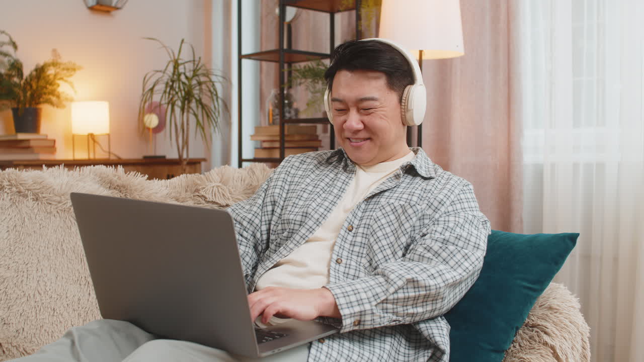 Happy relaxed overjoyed chinese middleaged man in wireless headphones listening to music on sofa