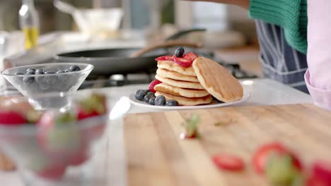 midsection of african american mother and daughter pouring honey on pancakes in kitchen, slow motion