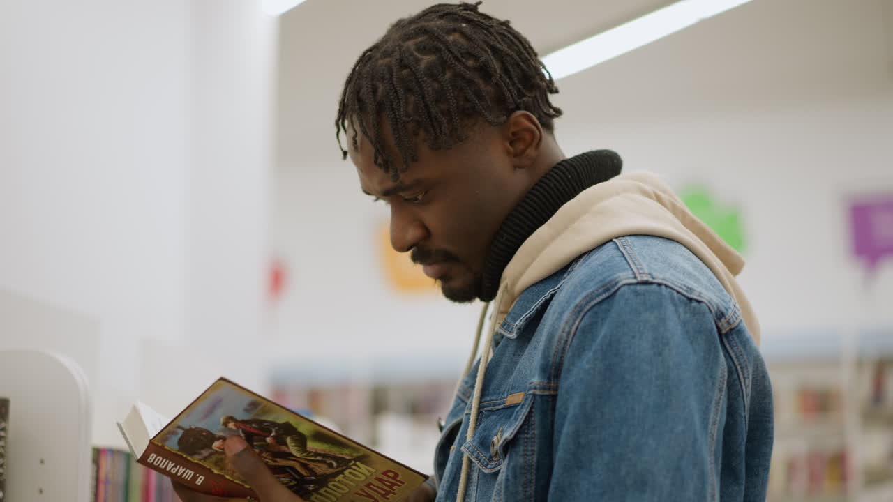Male in denim jacket focused on inspecting book in library. He examines pages with concentration in modern library, surrounded by colorful books on well-organized shelves in peaceful environment