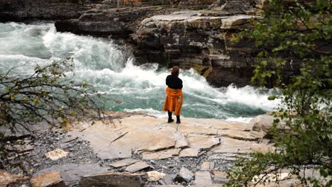 female hiker standing near ledge taking photo of raging flowing river water in sweden