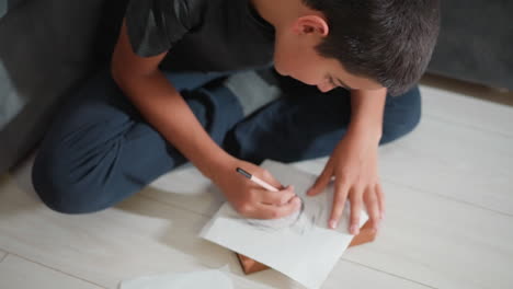 little boy seated cross-legged on floor deeply focused on sketching portrait with pencil during quiet creative time near sofa capturing early artistic learning and imagination in natural home setting