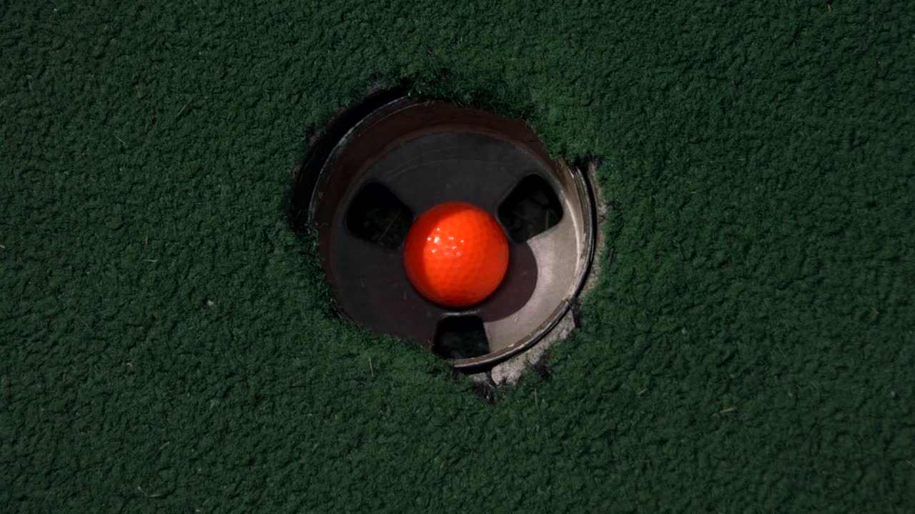 A close up birds eye view of an orange ball in a hole and then a purple and yellow golf ball fall into the golf hole on top of each other
