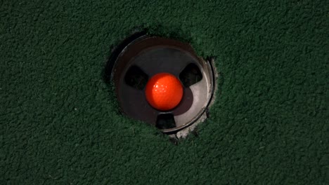 Eine-Nahaufnahme-Eines-Orangefarbenen-Balls-In-Einem-Loch-Aus-Der-Vogelperspektive,-Und-Dann-Fallen-Ein-Violetter-Und-Ein-Gelber-Golfball-übereinander-In-Das-Golfloch