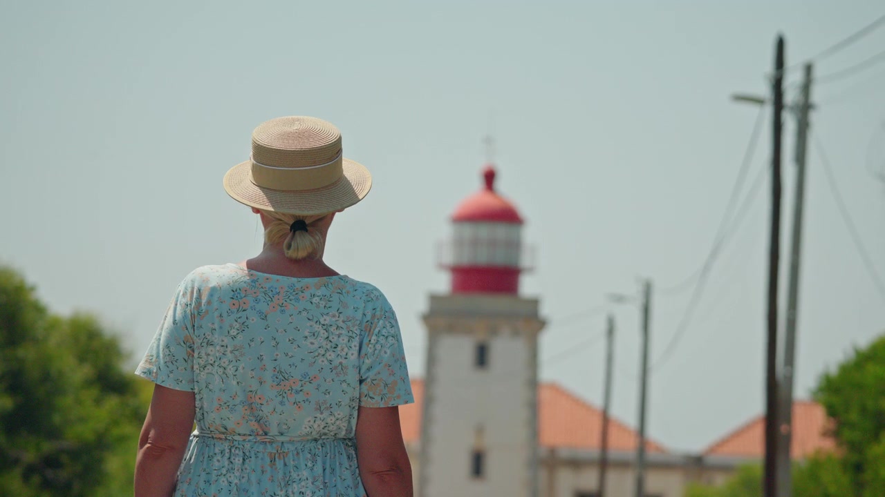 Donna in abito estivo e cappello che guarda un faro.