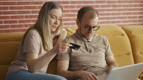Wife-and-husband-on-couch-together-making-purchase-online-on-laptop,-paying-with-credit-card