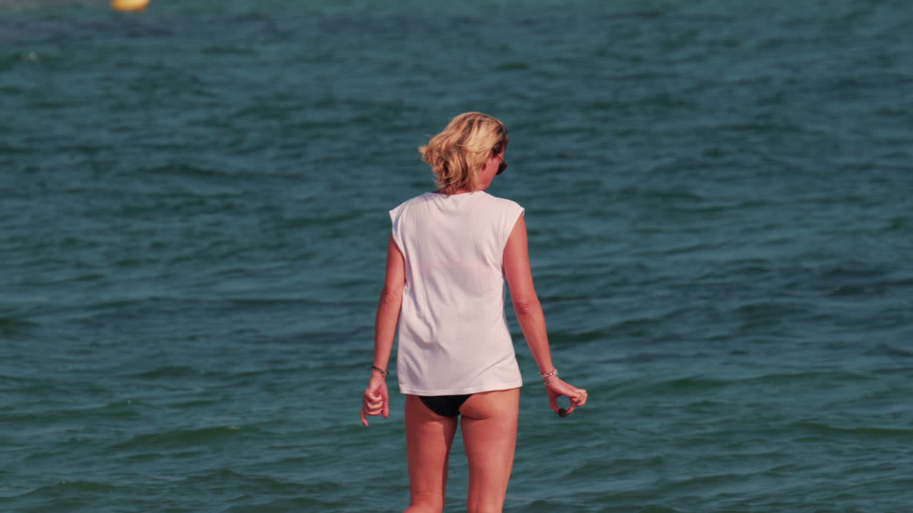 A woman with short blond hair stands waist deep in the sea, facing the horizon