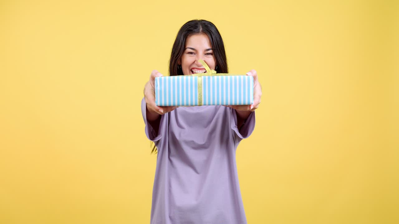 Young woman smiling and presenting a gift box on a yellow background