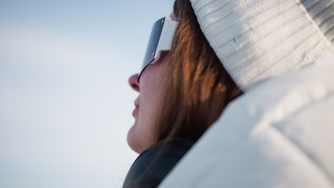 Side view of woman adjusting fancy tinted goggles under clear sky wearing white beanie and warm coat gentle breeze tousles hair sunshine reflects off visor conveying cheerful winter fashion mood