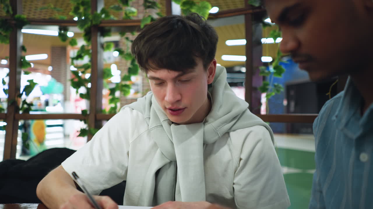 Man in blue shirt holds phone to show screen while friend writes notes on paper pad, scene set in bright office with soft natural light and blurred background conveying focused collaboration