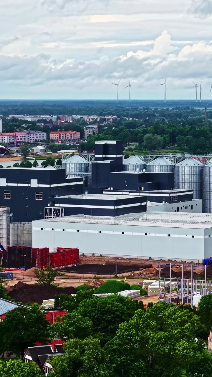 Stort industrianlæg med siloer og en fjern vindmøllepark under dramatisk skydække, vertikalt luftfoto mens regn falder på fabriksbyggeri.