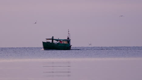 Fishing boat moves through calm pastel waters at sunrise, distant lights visible on the horizon
