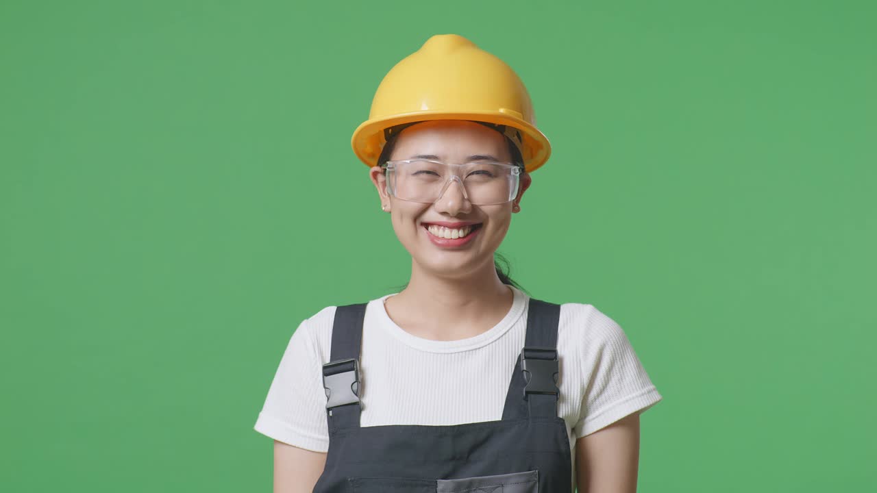 안경과 안전 헬을 착용 한 아시아 여성 노동자가 녹색 스크린 배경 스튜디오에서 카메라에 서서 미소 짓고 있습니다.