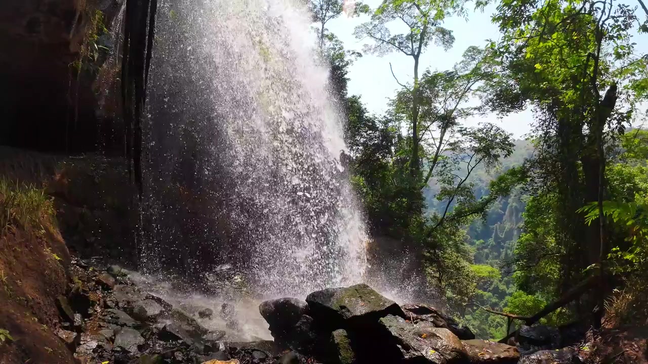 Tampilan gerak lambat dari balik Air Terjun Manchewe di Livingstonia, Malawi, saat air yang diterangi matahari menghantam bebatuan di samping lembah hijau cerah.