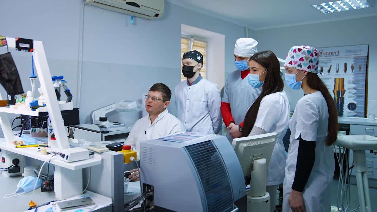 Group of students in masks stands behind the teacher looking on computer screen. Medic shows the denture x-ray to future dentists.