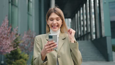 excited female user customer standing in urban street using smartphone, get amazing sale offer winning online bid, feel ecstatic reading great online news at outside