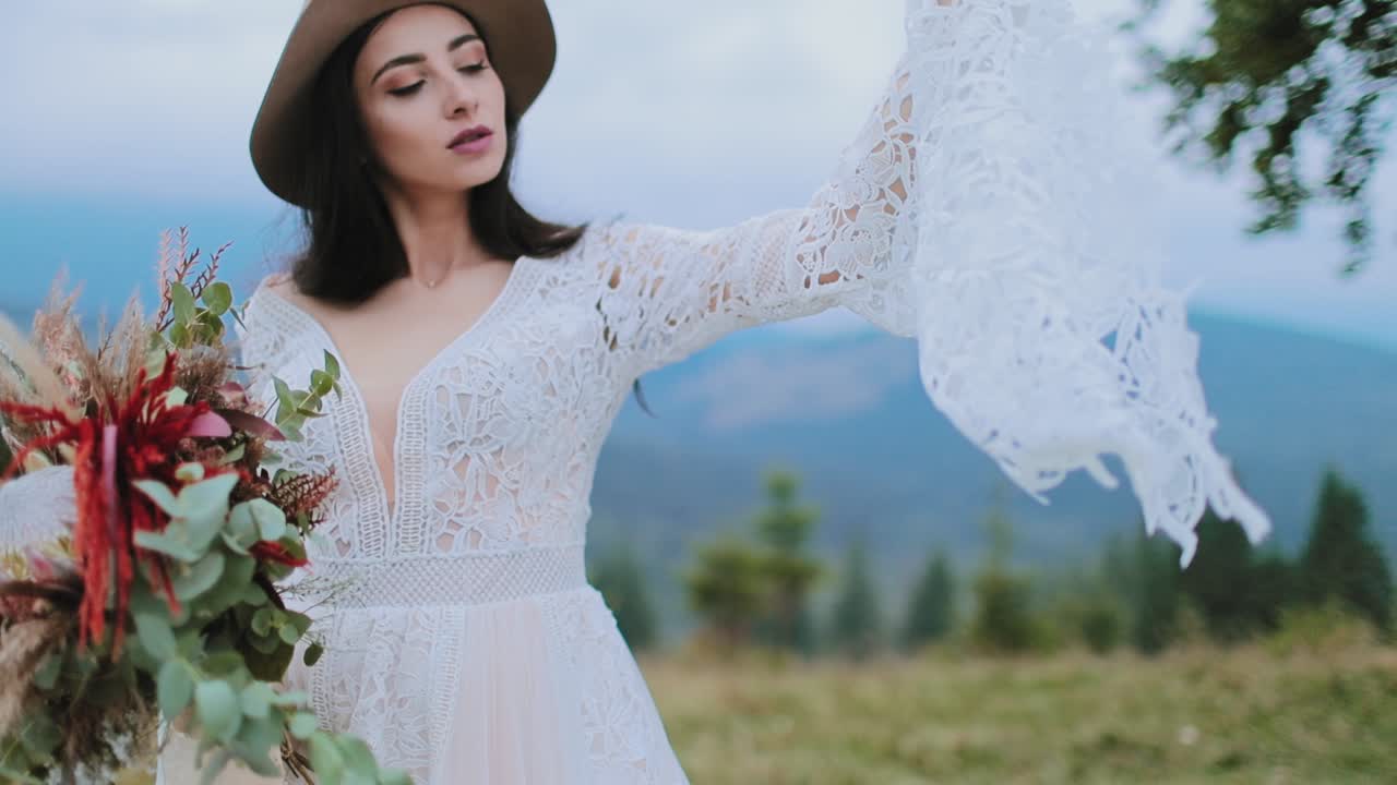 Beautiful woman in wedding dress with waving sleeves. Attractive bride in hat and white dress with flowers in hands against the mountains.