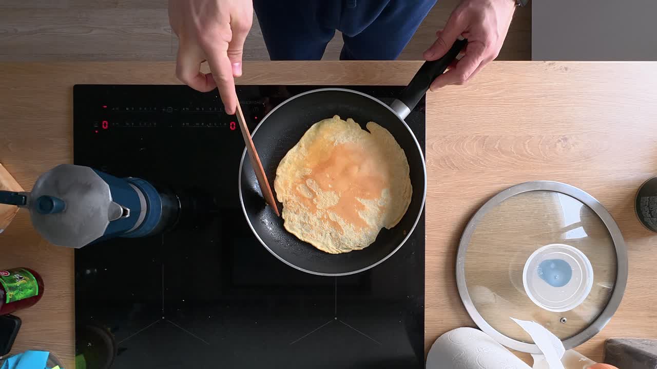 Overhead view of a person cooking a pancake on a modern stovetop. The batter is flipped with a spatula, highlighting detailed kitchen action and culinary skills