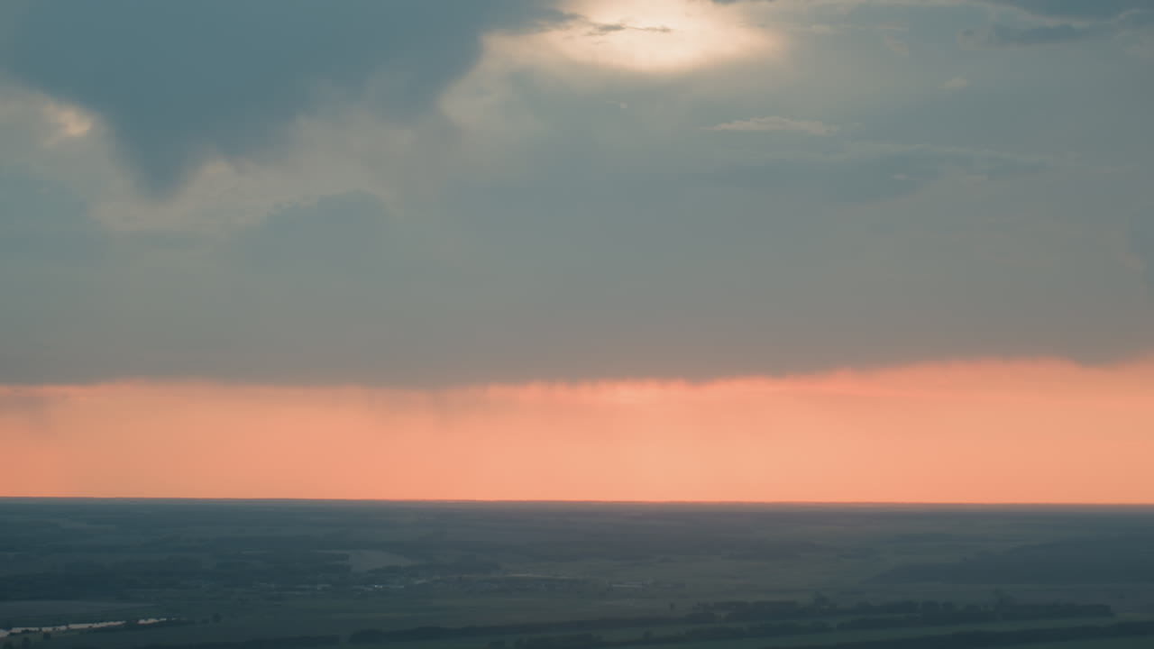 aerial sunset light filters through dramatic clouds over vast patchwork farmland and distant fields creating moody rural panorama with soft pastel sky subtle rain shafts on horizon