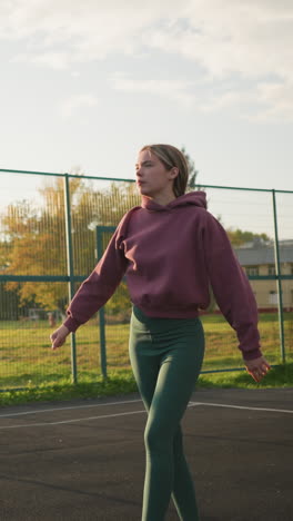 mujer atlética en leggings verdes lanza el voleibol para servir en la cancha al aire libre con campo abierto y persona caminando en el fondo, jugador enfocado preparándose para iniciar la acción del juego