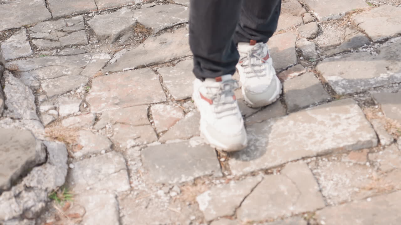 White sneakers walking on cobblestone pavement, closeup sequence of feet in white trainers and dark jeans moving across cracked historic stones, textured uneven surface, slow casual stride suggesting