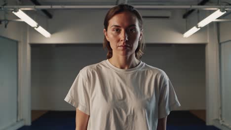 confident female athlete standing in shadowy gym, white t shirt highlighting muscular physique, preparing for intense training session with unwavering determination