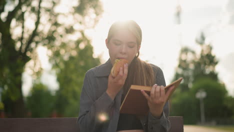 lady in grey top enjoying snack while reading book outdoors, seated on park bench, surrounded by trees and greenery with blurred background, soft sunlight illuminates