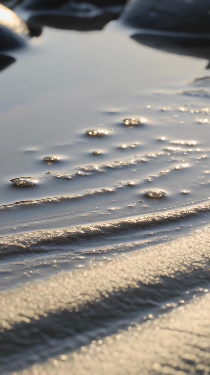 Video vertical: Mostrando el agua de una piscina reaccionando a un suave movimiento en la playa, con las crestas de arena ondulando y burbujeando.