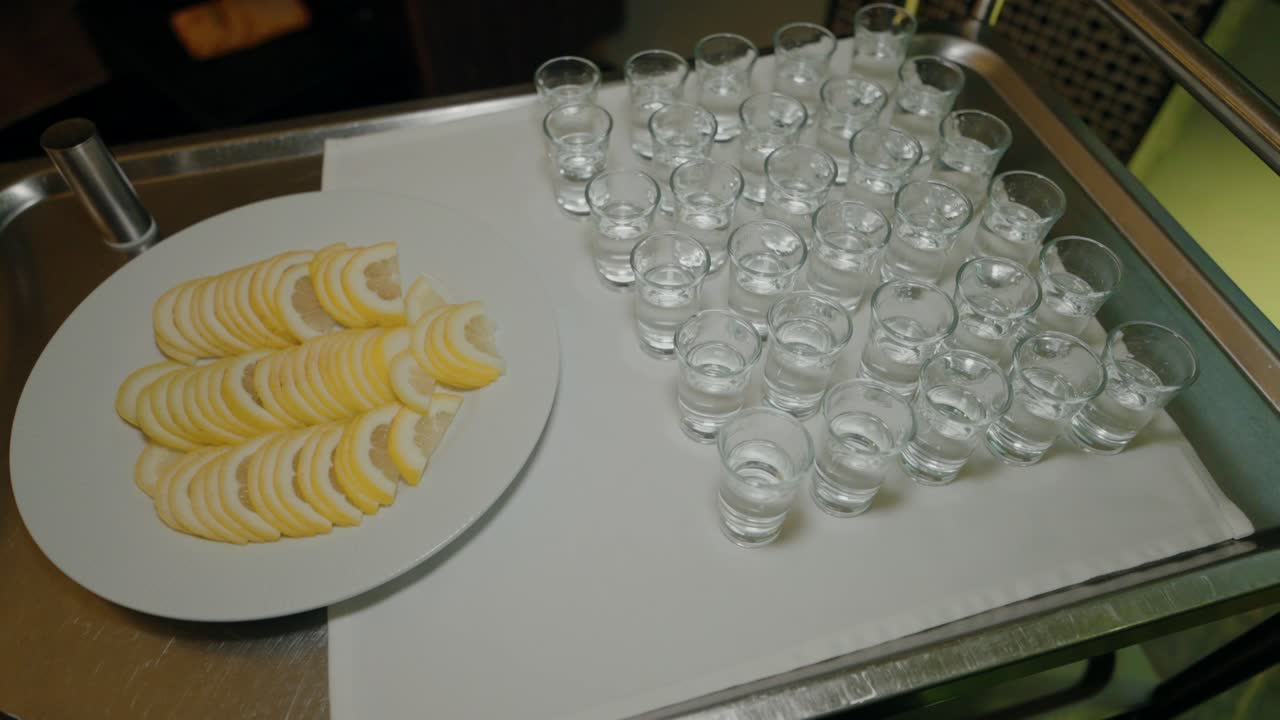 Tray of arranged lemon slices beside a set of empty shot glasses on a white cloth.