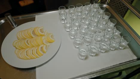 tray of arranged lemon slices beside a set of empty shot glasses on a white cloth.