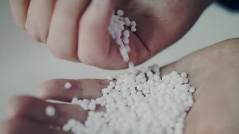 an extreme close-up shot of a person's hands inspecting white, spherical fertilizer granules. the pellets are poured from one hand to another, some falling in slow motion