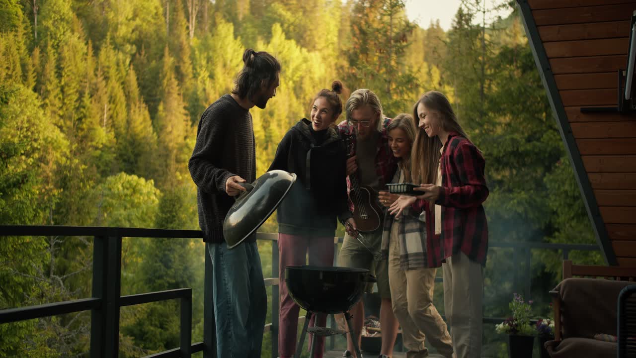 Five friends cook food on the grill on the balcony of a country house overlooking the mountains and green forest. Friends chatting and laughing at a picnic