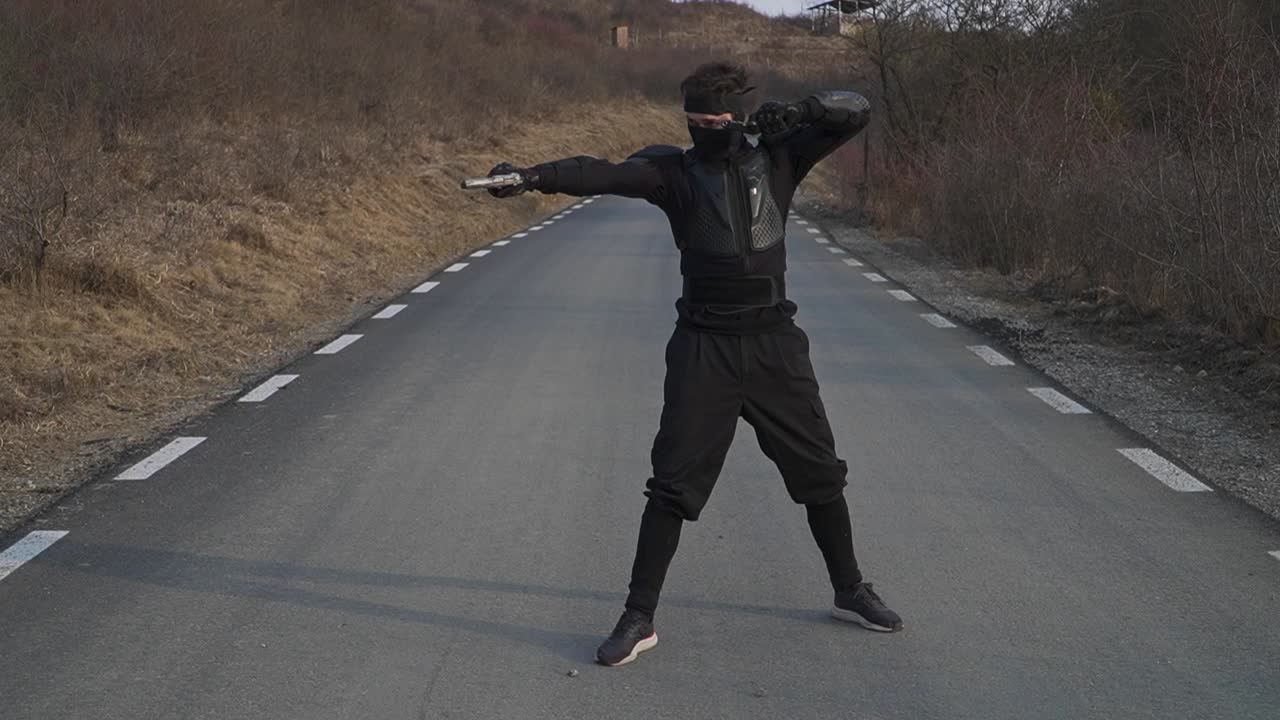 A figure in a black ninja costume strikes a pose with guns on a deserted asphalt road under a clear sky. Dramatic and action-oriented.