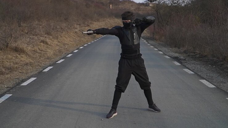 A figure in a black ninja costume strikes a pose with guns on a deserted asphalt road under a clear sky. Dramatic and action-oriented.