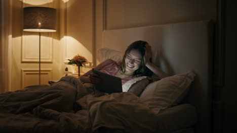 girl watching funny tablet video movie at night. happy woman resting home bed