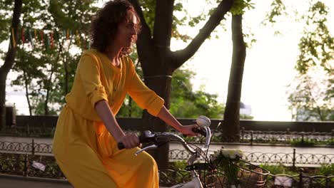 Attraktive-Frau-Im-Langen-Gelben-Kleid-Radelt-Im-Sommer-Auf-Dem-Fahrrad-Durch-Die-Stadt,-Mit-Korb-Und-Blumen-Im