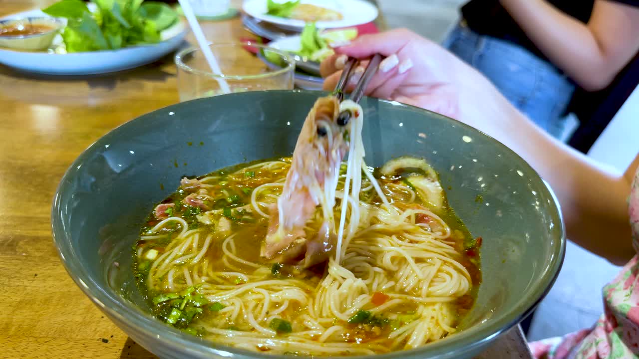 A person uses chopsticks to lift noodles from a flavorful Vietnamese soup, set in a casual dining environment